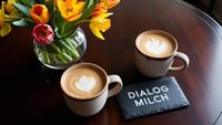 Zwei Tassen mit Milchcafés auf einem Holztisch mit einer Vase mit Frühlingsblumen und einer Schiefer-Tafel 